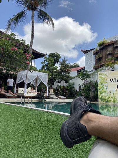 Resort style hostel, POV shot of person looking at pool, leg in frame, other people sitting by the pool, green lawn sunny skies